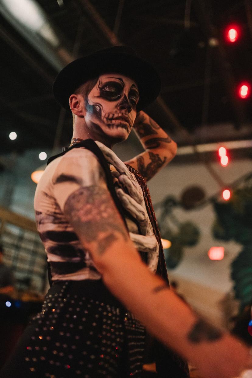 drag performer with skull face paint, reaching their left hand to hold their fedora hat