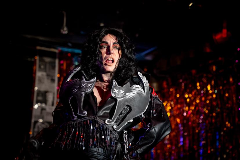 drag performer wearing a leather jacket with chains, sequins, and 2 black cats on the lapels. The artist has long black hair and one hand on hip