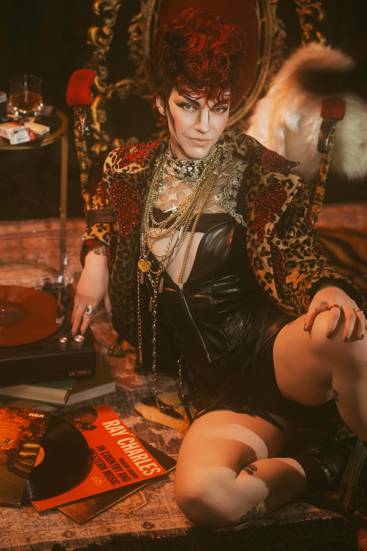 Drag king with red and black curly mohawk wig, wearing a leopard and rose print jacket, leather corset and shorts, and lots of gold chains and necklaces, sits back against a maroon and gold chair. A record player and a Ray Charles album is on the floor