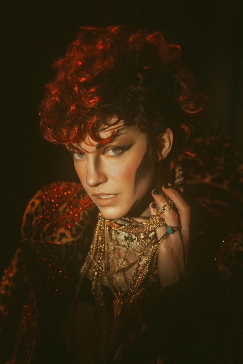 drag performer close up portrait, wearing a red and black curly mohawk wig and lots of gold jewelry, along with a leopard and rose print coat