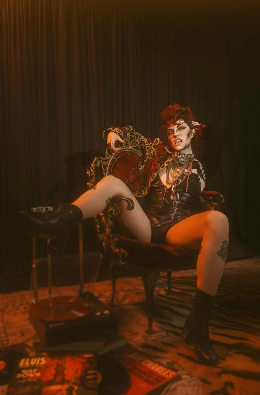 drag performer sitting in an ornate maroon and gold chair, wearing leather corset and shorts, along with a leopard and rose print coat and lots of gold jewelry